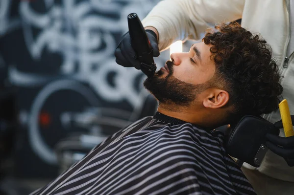 Modern, şık bir kuaför salonunda elektrikli traş makinesi kullanarak berberden karmaşık bir sakal kesimi alan adam.