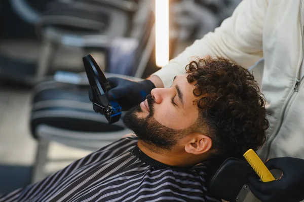 Genç bir adam, modern bir berber dükkanında elinde makasıyla bir berberden profesyonel saç kesimi ve sakal kesimi alıyor.