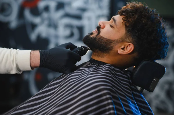 Berber koltuğunda oturan bir adam eldiven takan profesyonel bir berberden sakal tıraşı oluyor.