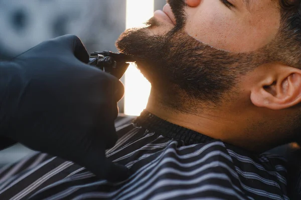 Siyah eldiven giyen bir berberden sakal tıraşı olan bir adam, kesin stil ve şekle odaklanıyor.