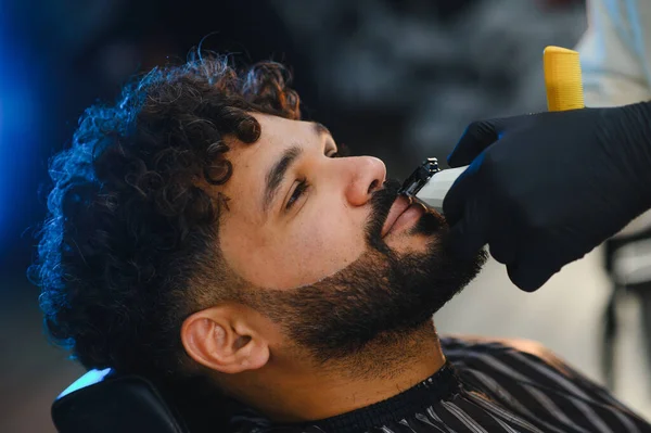 Kıvırcık saçlı, sakallı ve bıyıklı bir adam siyah eldivenli bir berber tarafından tıraş ediliyor.