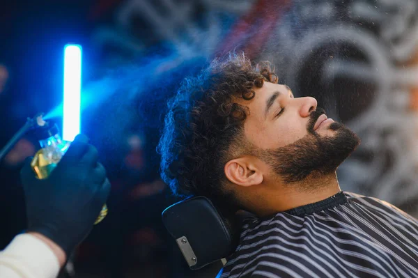 Berber dükkanı çalışanı parfüm sıkıyor ya da müşterinin sakalına saç spreyi sıkıyor. Bakım ve bakım için kıvırcık saçlı.