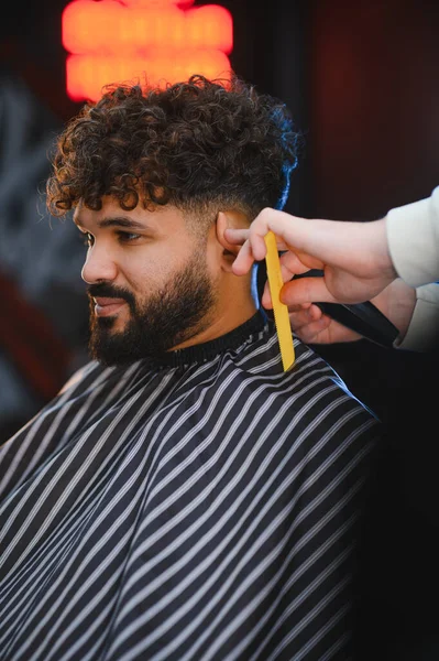 Kıvırcık saçlı ve sakallı bir adam profesyonel bir berber dükkanında tıraş makinesiyle yeni bir saç kesimi yaptırıyor.
