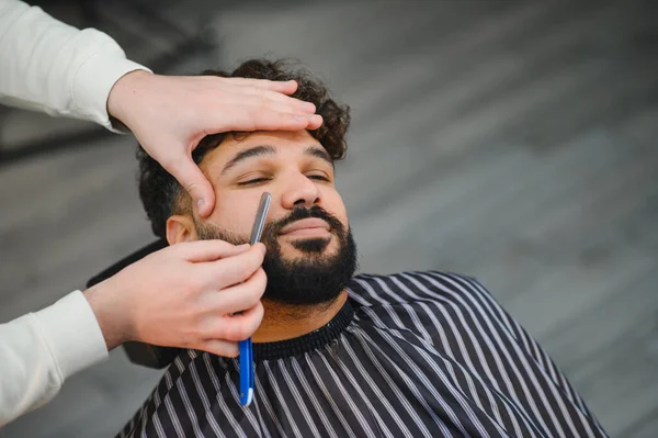 Berber bir adamın sakalını ustura ile dikkatlice şekillendirip buduyor, berber dükkanında profesyonel tımar hizmeti veriyor.