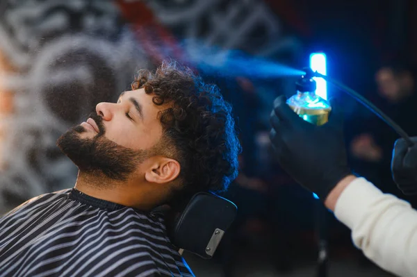 Kıvırcık saçlı ve sakallı bir adam modern bir kuaförde kuaförlük yaptığı için saçını spreyleyen bir berber ile eğleniyor.