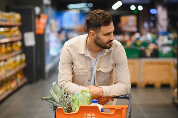 Ananas, marul ve süt gibi taze yiyeceklerle dolu turuncu bir alışveriş arabasını iyi aydınlatılmış bir süpermarkette iten adam.