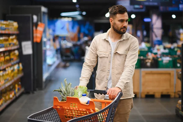 Parlak bir süpermarkette alışveriş yapan sakallı bir adam, dolu bir arabayı iterken düşünceli bir şekilde bakkaliye ve taze ürünleri seçiyor.