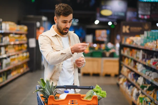 Adam uzun bir süpermarket faturasını kontrol ediyor, market harcamalarının artması ve bütçe düzenlemesi konusunda şok ve endişe gösteriyor.