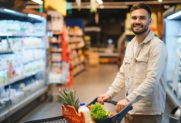 Gülümseyen, alışveriş arabasını iten genç bir adam. Parlak bir süpermarket koridorunda içi taze yiyeceklerle dolu.