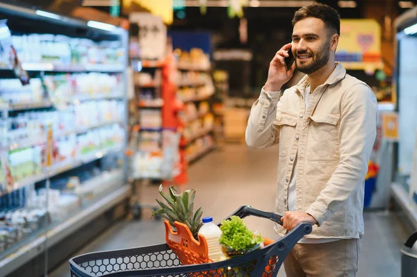 Market koridorunda bir telefon görüşmesi yaparken alışveriş arabasını iten adam. Modern tüketici alışkanlıklarına odaklan