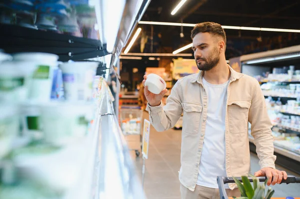 Genç adam bakkal alışverişi yapıyor, bir marketin parlak ışıklı süt ürünleri bölümünde paketlenmiş yoğurt kutularını karşılaştırıyor.