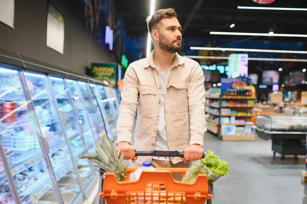 Genç bir adam taze ürünlerle dolu turuncu bir alışveriş arabasını itiyor. Dondurucudan bir şeyler seçiyor.