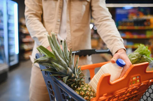 Bir adamın elleri marketin koridorunda içi taze ürün, süt ve diğer şeylerle dolu bir alışveriş arabasını itiyor.