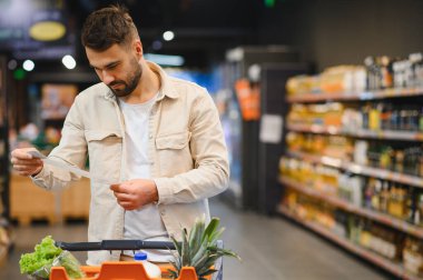 Market koridorunda alışveriş listesi üzerinde çalışan, akıllı alım ve bütçeye odaklanan bir adam.