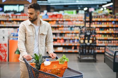 Genç adam markette içi taze ürün, süt ve diğer şeylerle dolu bir alışveriş arabasını itiyor.