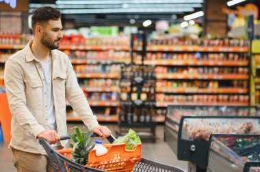 Genç adam taze ürünler ve temel ihtiyaçlarla dolu bir arabayı itiyor. Parlak bir süpermarket koridorunda geziniyor.