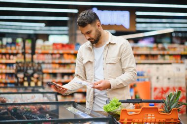 Süpermarketteki dondurucuda dondurulmuş ürünleri karşılaştıran bir adam taze ürünlerle dolu bir arabayı iterken market alışverişine odaklanıyor.