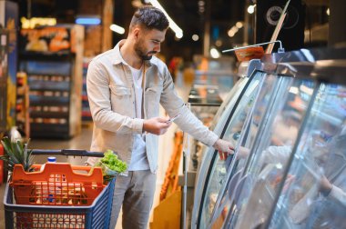 Market alışverişi yapan bir adam, parlak bir süpermarket koridorunda soğutmalı vitrinde ürünleri kontrol eden bir adam.