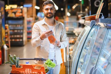 Bir markette paketlenmiş somon filetosu tutan bir adam alışveriş arabası için sağlıklı yiyecek seçiyor.
