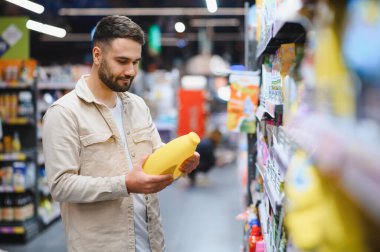 Bir mağaza koridorunda sarı bir çamaşır deterjanı şişesini inceleyen bir adam ev ürünleri için satın alma kararı veriyor.