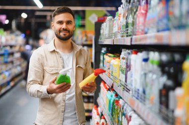 Süpermarketteki genç adam iki şişe temizlik malzemesini karşılaştırıyor, hangi ev ürününü alacağına karar veriyor.