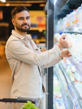 Süpermarketteki soğutuculu raftan taze süt seçen bir adam. Süt ürünleri, sağlıklı yiyecekler ve bakkaliye alışverişi.