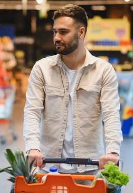 Genç bir adam markette sağlıklı gıda seçenekleri arayan taze ürünlerle dolu bir alışveriş arabasını itiyor.