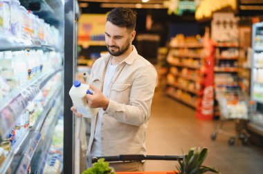 Odaklanmış bir adam canlı bir süpermarkette soğutuculu bölümden bir şişe süt seçiyor.