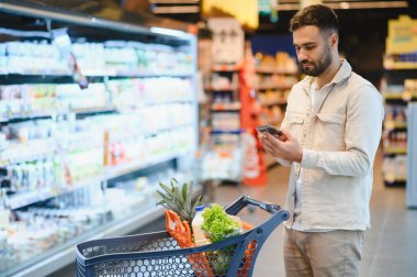 Markette alışveriş yapan bir adam, el arabasındaki eşyaları cep telefonundaki listeyle karşılaştırıyor. Modern tüketim kavramı