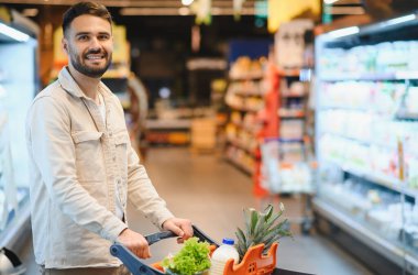Gülümseyen bir adam, markette taze sebze ve meyve gibi sağlıklı yiyecek malzemeleriyle dolu bir alışveriş arabasını iterken.