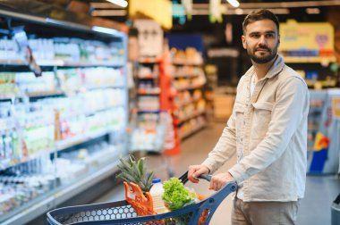 Genç adam taze ürünler, süt ve diğer şeylerle dolu bir alışveriş arabasını parlak bir markette itiyor.