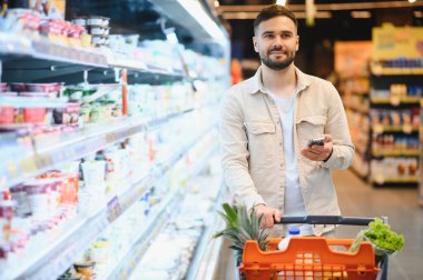 Market koridorunda alışveriş arabalarını iten genç adam raflara bakıyor ve akıllı telefondan alışveriş listesini kontrol ediyor.