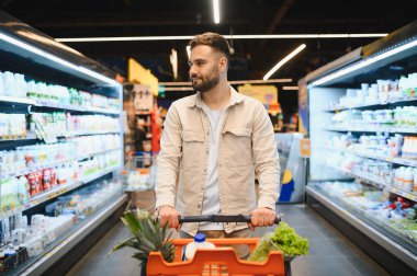 Taze sebze ve sebzelerle dolu bir alışveriş arabasını iten adam parlak ışıklı bir süpermarket koridorunda yürüyor.