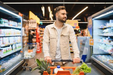 Markette koridorları gezerken içi taze yiyecek dolu alışveriş arabalarını iten adam. Modern yaşam tarzı ve perakende konsepti