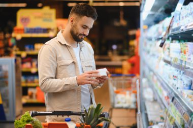 Elinde küçük bir paket tutan adam, etiketi dikkatle okuyor ve market alışverişi yaparken beslenme bilgilerini okuyor.