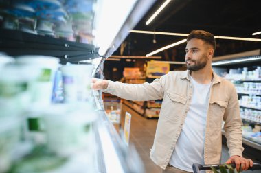 İnsan, modern süpermarkette yoğurt seçerek soğutulmuş koridora uzanır ve seçenekler arasından taze süt ürünlerini seçmeye odaklanır.