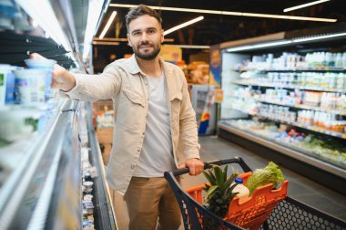 Gülümseyen adam buzdolabından süt ürünlerini seçerken bir alışveriş arabasını koridorda itiyor.