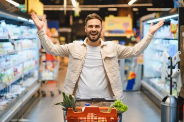 Neşeli erkek müşteri market alışverişi sırasında iyi anlaşmaları kutluyor, koridorda dolu bir el arabasını itiyor.