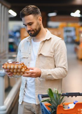 Adam elinde yumurta kartonuyla markette taze ürün ararken içindekileri inceliyor.