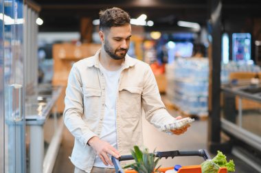 Modern bir markette içi taze ürünlerle dolu bir alışveriş arabasını iterken elinde bir paket yumurta tutan adam.