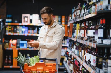 Mağaza raflarındaki geniş bir seçimden alkollü içecekleri karşılaştıran ve seçen erkek tüketiciler, bir alışveriş arabasını iterek