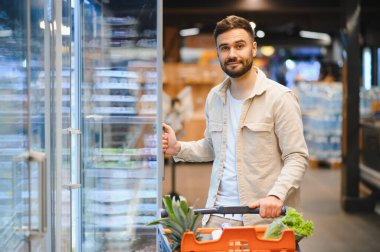 Markette alışveriş yapan genç bir adam, elinde dolu bir el arabası, cam bir buzdolabı kapısı açıp ürün arıyor.