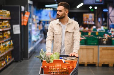 Taze ürünlerle dolu alışveriş arabasını iten adam, bakkal koridorunda eşyalar arıyor. Tüketici yaşam biçimi