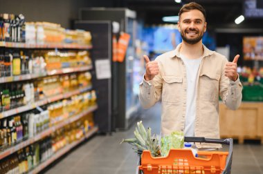 Mutlu sakallı adam bir süpermarket arabasını koridorda itiyor, geniş bir gülümseme içinde ve iki hevesli baş parmağını kaldırıyor.