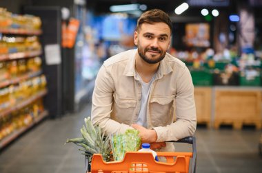Genç adam gülümsüyor, alışveriş arabasına yaslanıyor. Taze ürünlerle dolu, sağlıklı yaşam tarzı ve tüketicilik gösteriyor.