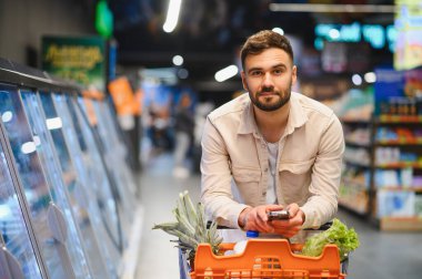 Market koridorunda alışveriş arabalarını iterken akıllı telefon tutan adam alışveriş listesine odaklanmış.