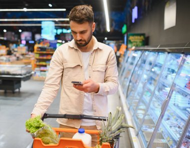 Alışveriş yapan kişi süpermarket koridorlarında geziniyor, ürünleri karşılaştırıyor ve bir cep telefonu uygulamasında alışveriş listesini kontrol ediyor.