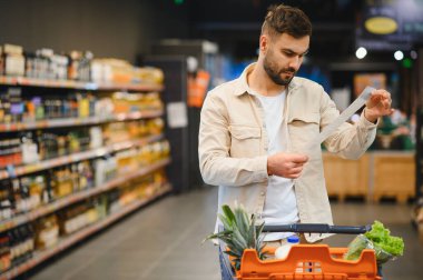 Alışveriş yaparken uzun bir satış makbuzunu inceleyen bir adam, yanında erzak taşıyan bir alışveriş arabası, masraflara ve bütçeye odaklanan biri.