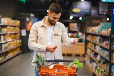 Genç bir adam alışveriş arabasını itiyor, alışveriş listesini kontrol ediyor ve markette sağlıklı beslenmek için ürünler seçiyor.