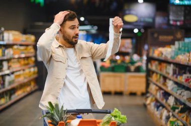 Elinde uzun bir makbuz tutan adam, yüksek masraflar ve market alışverişi sırasında artan gıda fiyatları karşısında şok olmuş görünüyor.